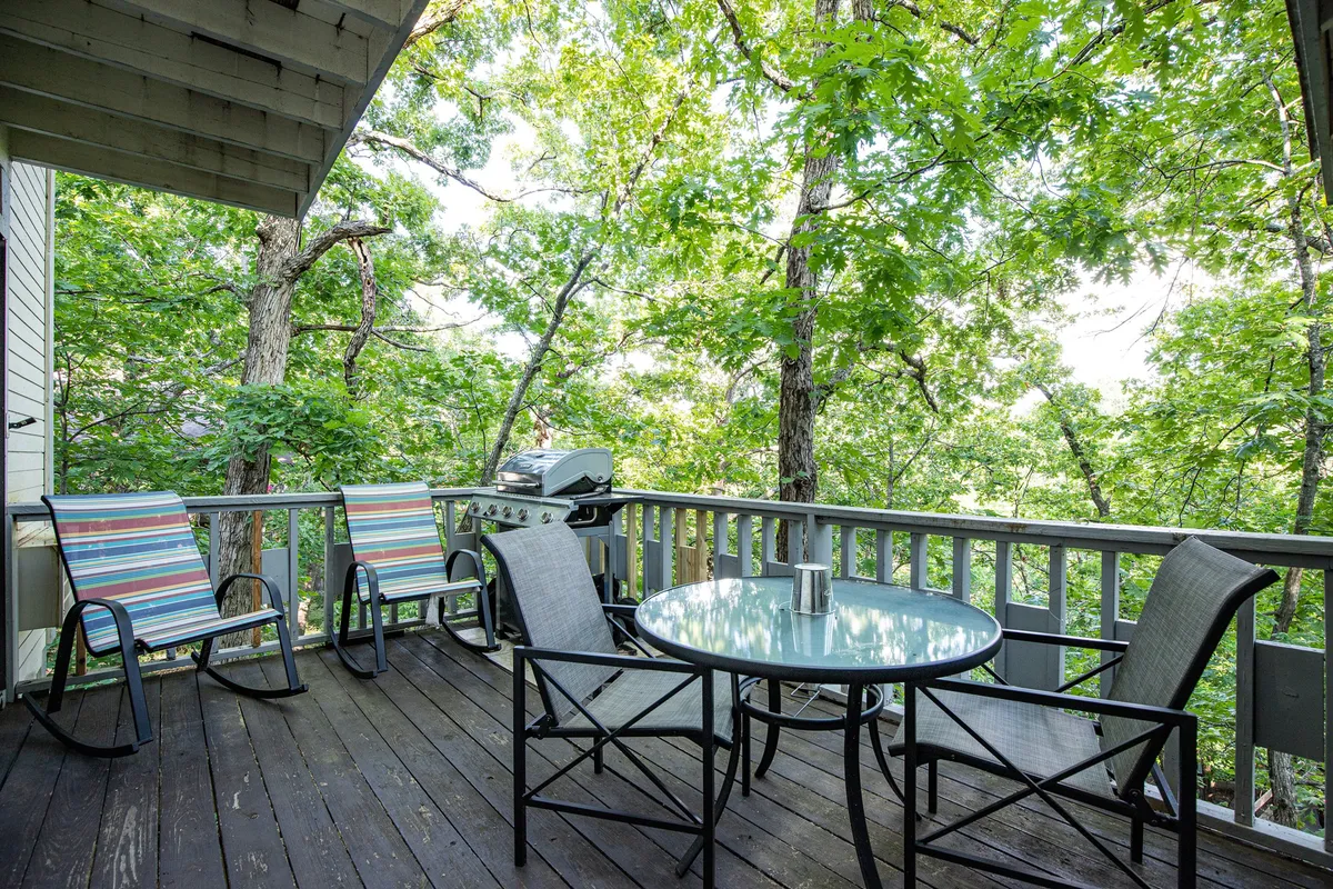 An inviting covered patio with a glass-top table, colorful chairs, a grill, and views of surrounding trees. The outdoor setting is perfect for al fresco dining, barbecues, and relaxed social gatherings in any season.