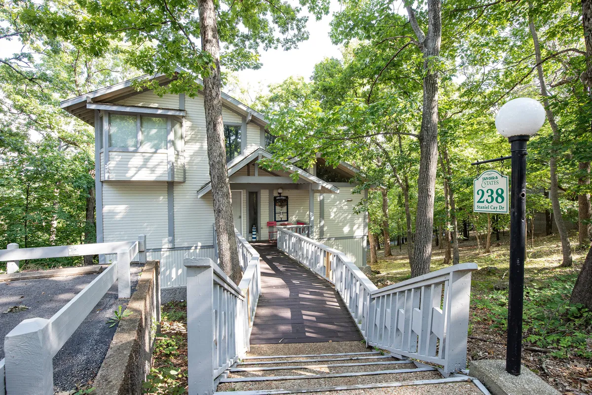 This exterior image shows the front entrance of the Cabernet Chalet, with a welcoming pathway leading up to the house, surrounded by lush trees. The inviting porch hints at spaces for relaxing or gathering outdoors.