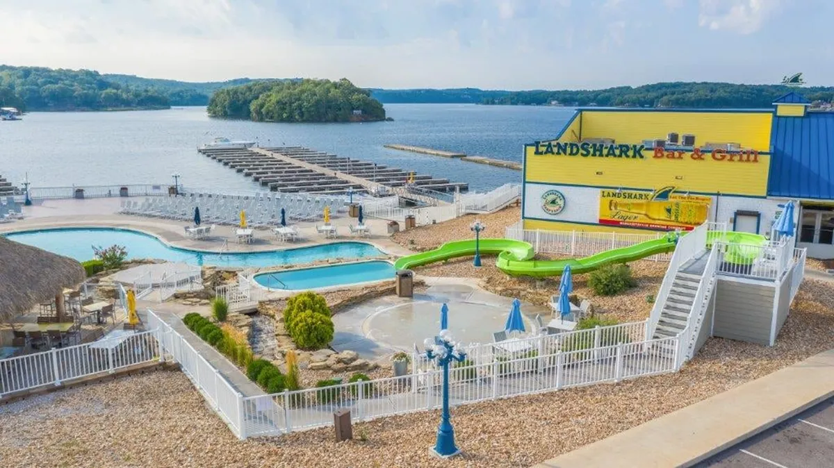 This vibrant exterior image captures a lively resort pool area featuring a swimming pool, a water slide, a bar and grill, and a picturesque lake view with a dock. The atmosphere is bustling, with guests swimming, dining, boating, and relaxing by the pool, all set against the backdrop of a glowing sunset.