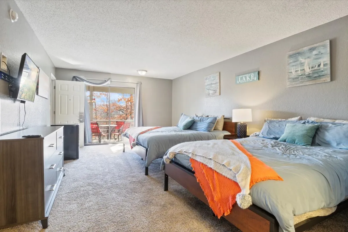 This image features two beds, a large window providing a serene view, a dresser, and a television. The room is bathed in natural light, creating a comfortable and inviting atmosphere that highlights the restful sleeping area and the connection to the outdoors.