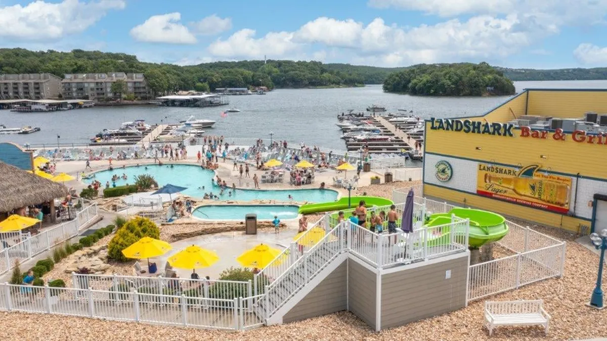 A lively resort-style pool scene with swimming pools, twisting water slides, a bar and grill building, and people enjoying various activities like swimming, sunbathing, dining, and socializing.