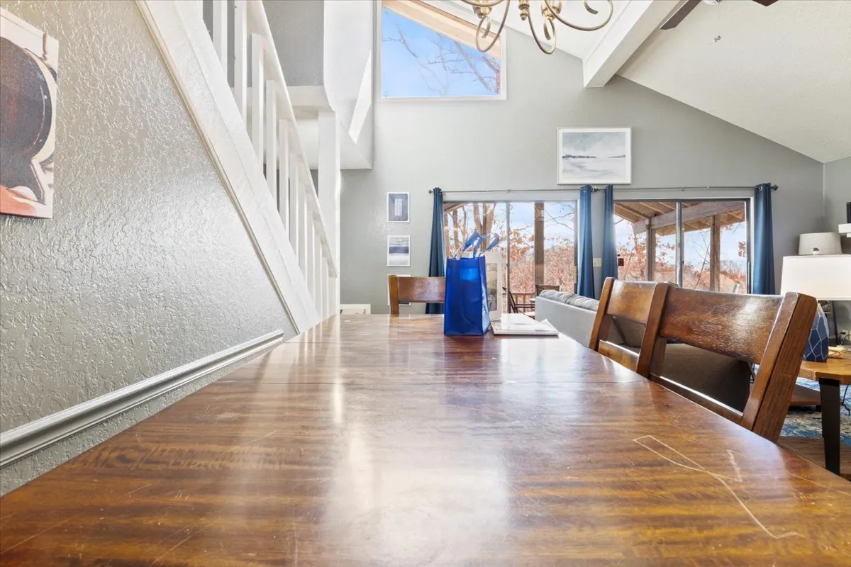 A long wooden dining table is the focal point of this warm, light-filled interior. The space is accented by a chandelier and large windows, creating an inviting setting for family gatherings, shared meals, or even working from home. A blue bag hints at the convenience and organization brought by guest services like the Pack Less Program.