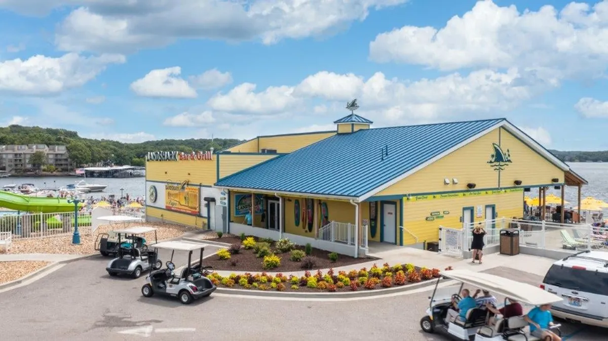 A picturesque lakeside restaurant with a view of the lake, golf carts parked outside, colorful landscaping, and a building that hints at activity and community. The setting suggests opportunities for outdoor dining, special events, and water or golf activities nearby.