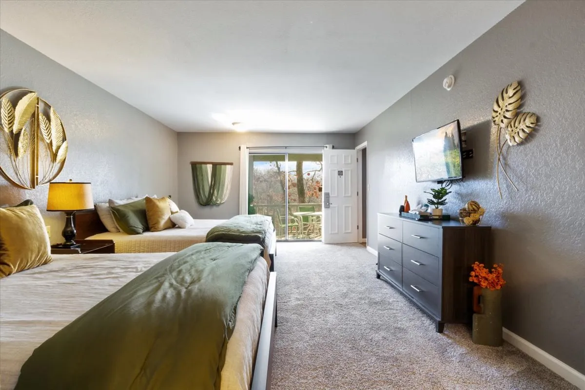 A welcoming suite with two beds dressed in green and beige bedding, decorative wall art, a dresser with stylish decor, and a large window leading out to a balcony. The setting is bright, modern, and provides a direct connection to the outdoors.