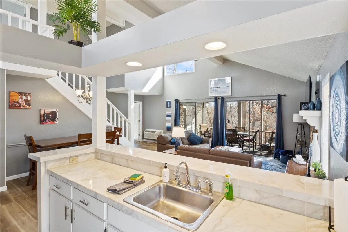 A stylish, open-plan kitchen and dining area with a table set for meals, a spacious kitchen sink, and comfortable living space in the background. Large windows flood the area with natural light, while the decor adds a welcoming, homey feel.