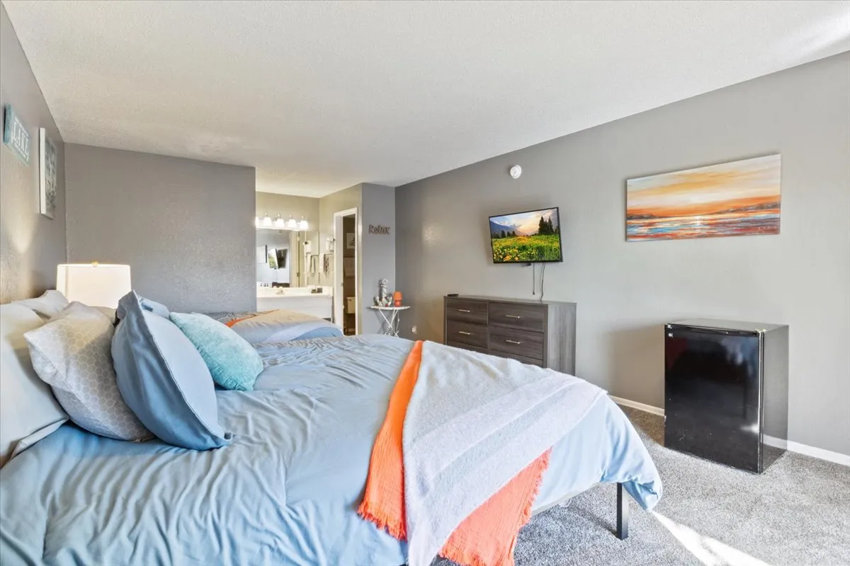 This cozy suite features a bed with colorful bedding, a TV mounted on the wall, a dresser adorned with decorative items, and a mini fridge. The setting feels personal, comfortable, and thoughtfully equipped for a restful stay.