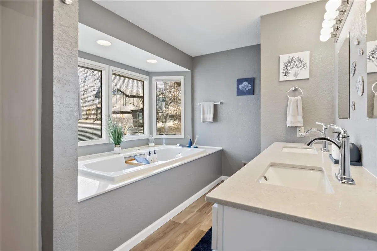 The image showcases a luxurious bathroom with a standalone bathtub, double sinks, large windows that let in abundant natural light, and tasteful decorative artwork. The space offers a tranquil setting ideal for relaxation and pampering routines.