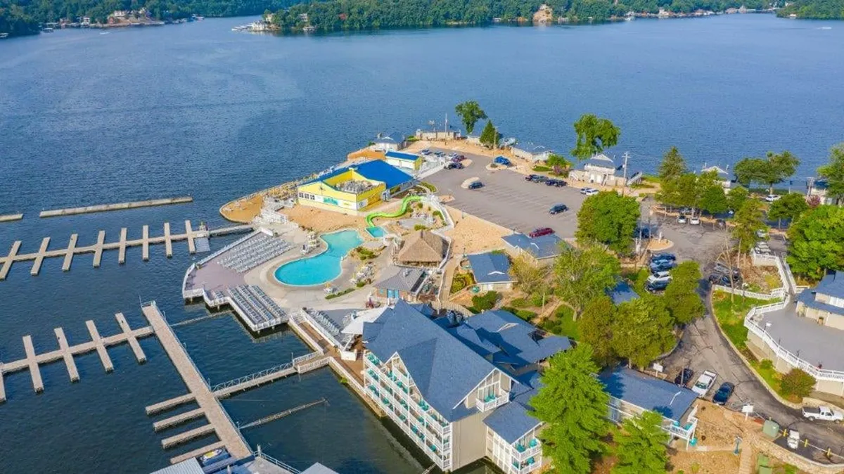 A panoramic outdoor view featuring a lake, swimming pool, dock, a colorful building, and a parking lot. The scene is bustling yet relaxed, offering multiple options for fun: swimming, boating, sunbathing, and social gatherings.